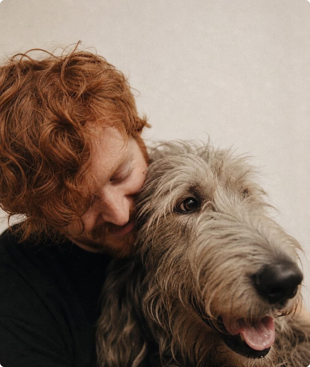 A young man with red hair closes his eyes and nuzzles his Irish wolfhound, who looks toward the camera.