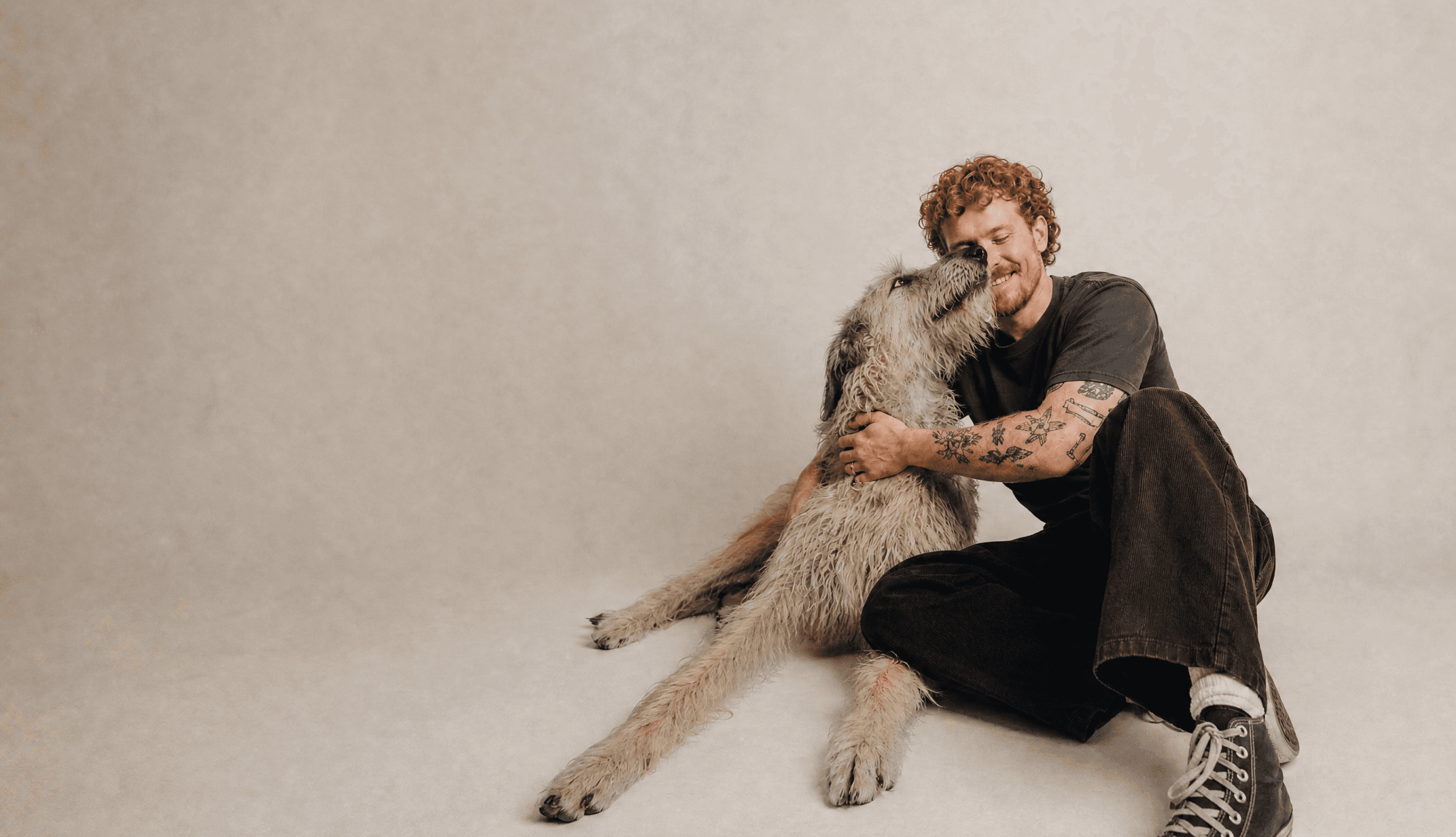 A young man sits on a studio floor hugging his Irish wolfhound, who leans in to nuzzle his face.