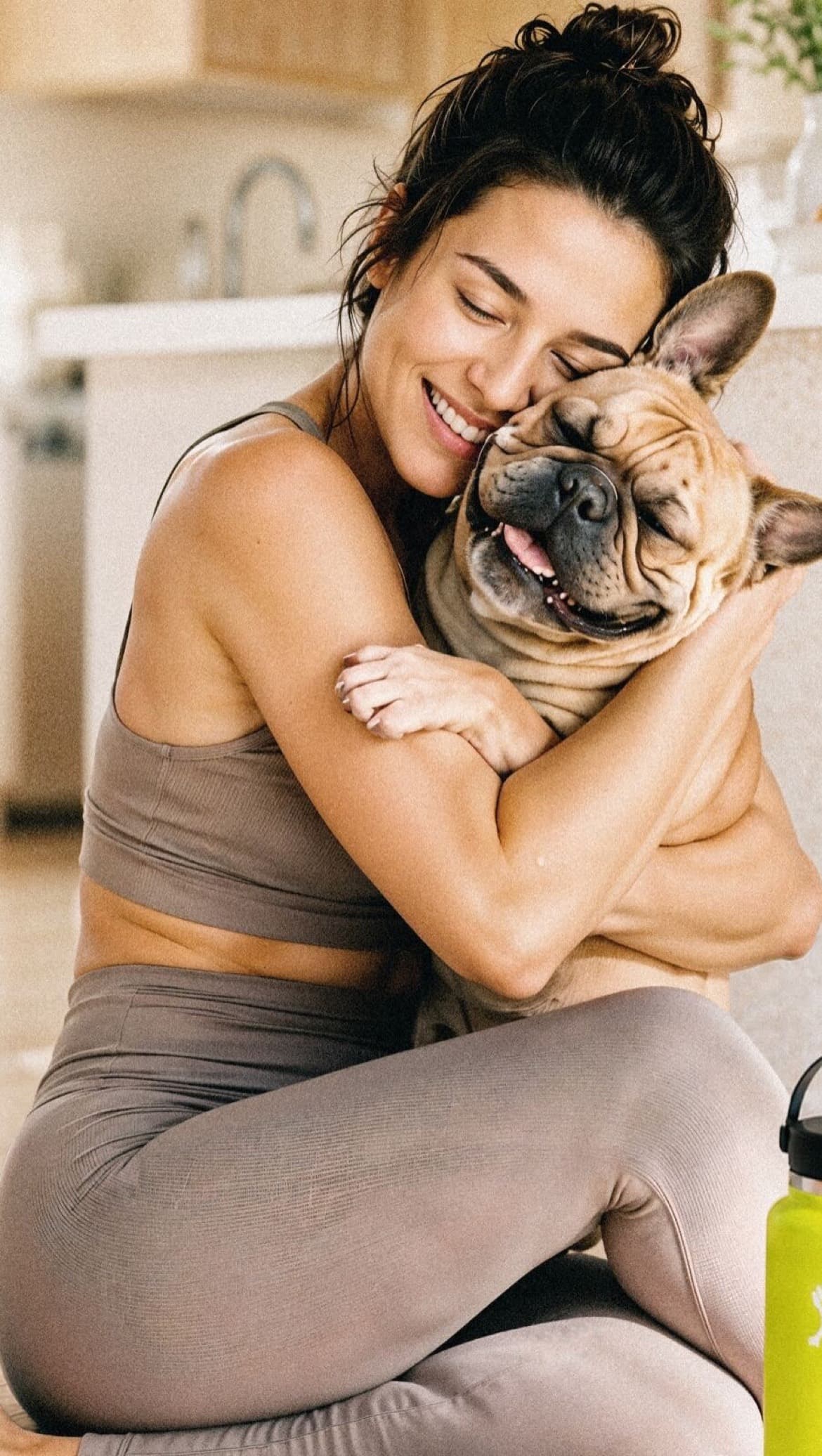 A woman sitting on a yoga mat hugs her smiling French Bulldog, both with their eyes closed.